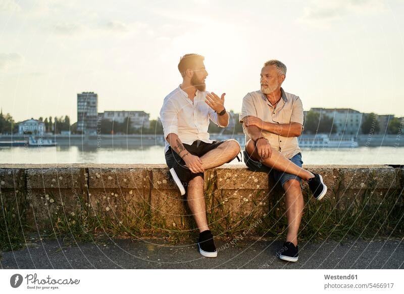 Vater und erwachsener Sohn sitzen auf einer Mauer am Flussufer und unterhalten sich Leute Menschen People Person Personen Europäisch Kaukasier kaukasisch 2