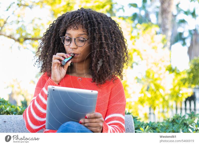 Afro Frau raucht elektronische Zigarette, während mit digitalen Tablette im öffentlichen Park Farbaufnahme Farbe Farbfoto Farbphoto Außenaufnahme außen draußen