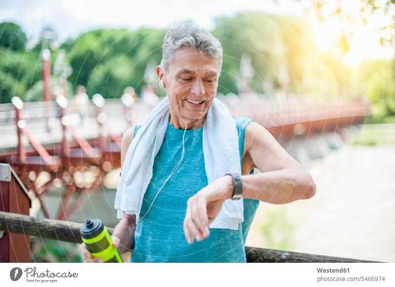 Lächelnder aktiver älterer Mann schaut auf intelligente Uhr, während er im Park steht Farbaufnahme Farbe Farbfoto Farbphoto Außenaufnahme außen draußen