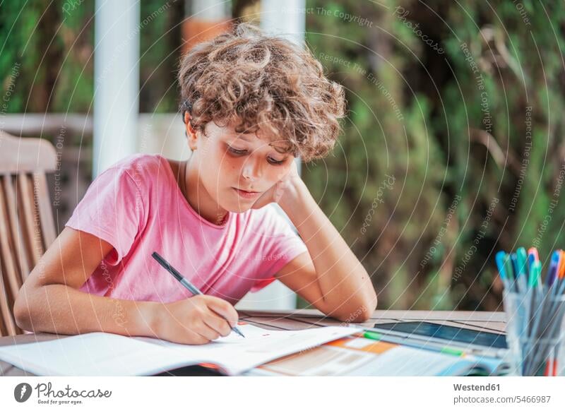 Junge sitzt am Gartentisch und macht Hausaufgaben Ausbildung Schueler Schulkinder Schüler Leute Menschen People Person Personen gelockt gelockte Haare