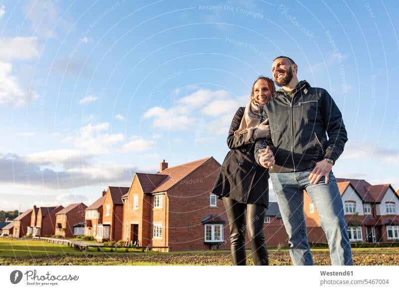 Glückliches Paar schaut weg, während es vor neu gebauten Häusern gegen den Himmel steht Farbaufnahme Farbe Farbfoto Farbphoto Tag Tageslichtaufnahme