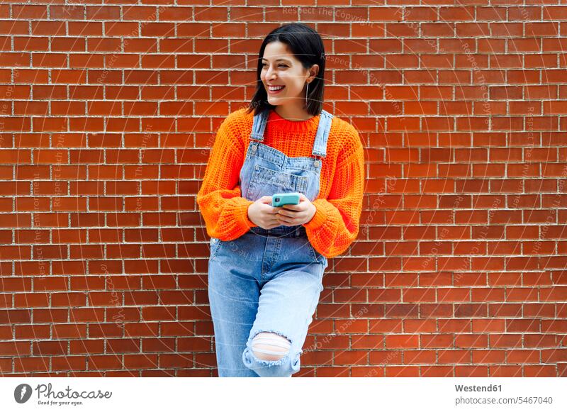Lächelnde Frau schaut weg, während sie an einer Ziegelmauer steht Farbaufnahme Farbe Farbfoto Farbphoto Außenaufnahme außen draußen im Freien Tag