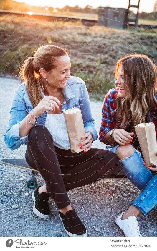 Zwei lachende Freunde sitzen auf Longboard und essen Popcorn Freundinnen Puffmais essend sitzend sitzt Freundschaft Kameradschaft Süßigkeiten Süßes Essen Food