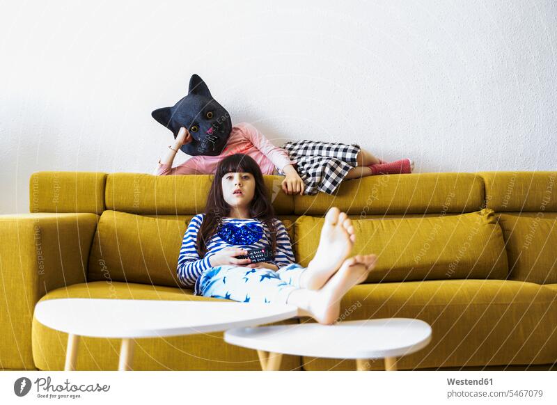 Zwei gelangweilte Mädchen sitzen auf der Couch, sehen fern, eine trägt eine Katzenmaske Couches Liege Sofas Fernbedienungen langweilig Farben Farbtoene Farbton