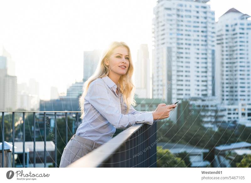 Blonde lächelnde Geschäftsfrau lehnt sich an ein Geländer und hält ein Smartphone auf dem Dach der Stadt Geschäftsfrauen Businesswomen Businessfrauen