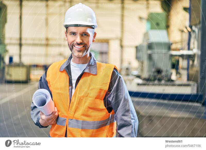 Porträt eines lächelnden Mannes mit Blaupausen in der Fabrik Portrait Porträts Portraits halten Fabrikgebäude Fabrikgebaeude Fabriken Männer männlich Bauplan