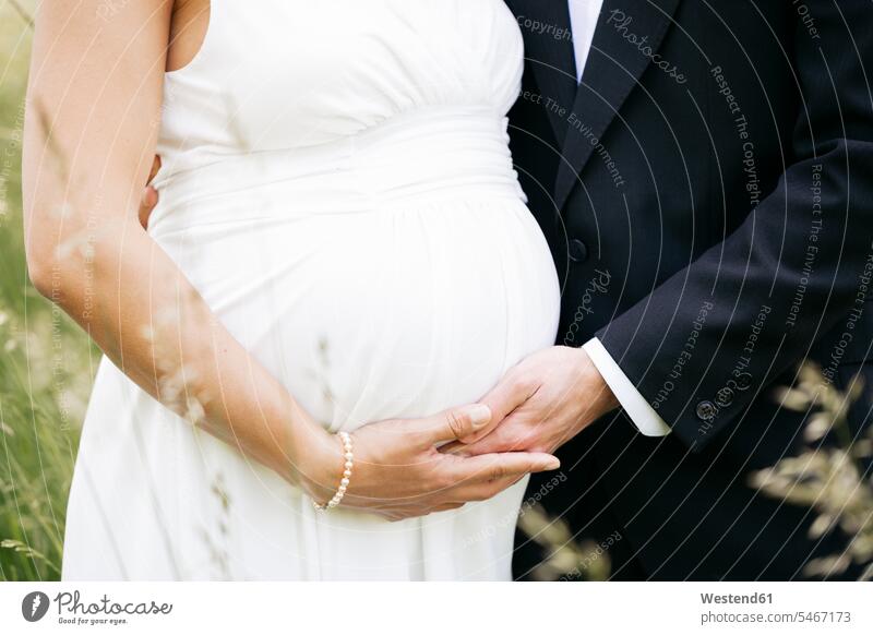 Schwangere Braut mit ihrem Ehemann hält Babybauch auf einer Wiese Leute Menschen People Person Personen verheiratet Ehepaare Ehemänner Ehefrauen Heirat heiraten
