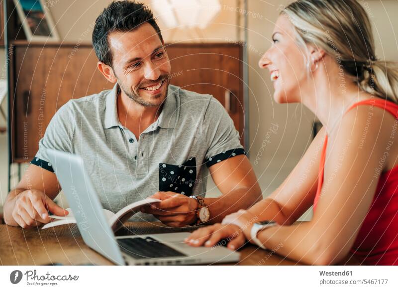 Glückliches Paar sitzt am Tisch mit Laptop und Buch Bücher Tische Holztische Rechner Laptops Notebook Notebooks Arbeit sitzend reden freuen Frohsinn