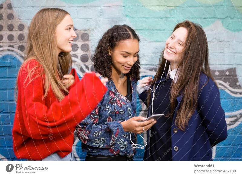 Drei Freunde stehen vor der Graffiti-Wand und hören mit Smartphone und Kopfhörern Musik Graffitis Freundinnen Ohrhörer hoeren iPhone Smartphones Freundschaft