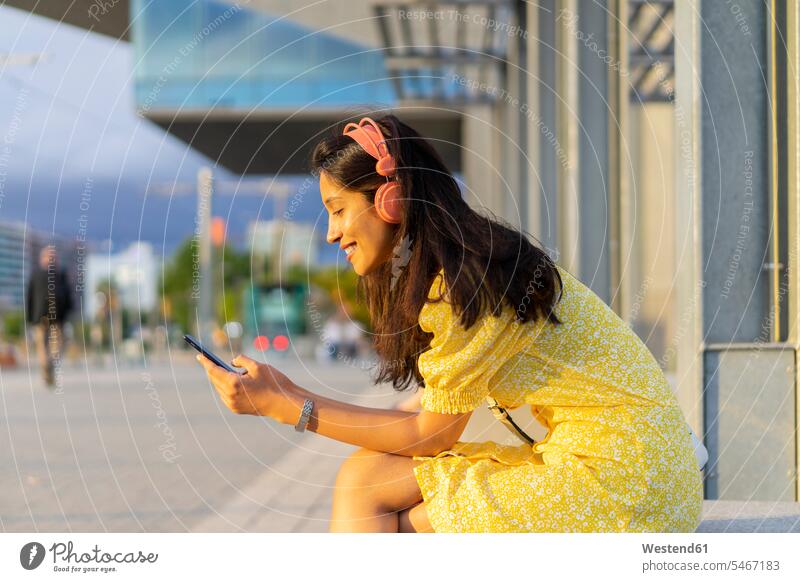 Glückliche Frau benutzt Smartphone und hört Musik, während sie in der Stadt sitzt Farbaufnahme Farbe Farbfoto Farbphoto Außenaufnahme außen draußen im Freien