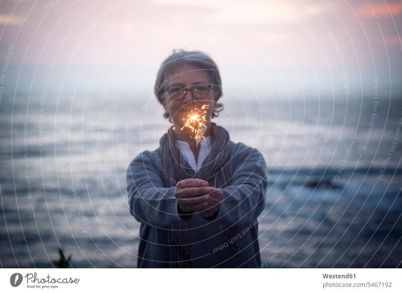 Glückliche ältere Frau steht vor dem Meer bei Sonnenuntergang hält Wunderkerze Spanien Stimmungsvoller Himmel romantischer Himmel Wunderkerzen Funke Funken