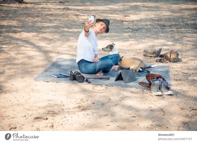 Frau sitzt auf einer Decke am Strand mit Hund und macht ein Selfie Decken weiblich Frauen Selfies Beach Straende Strände Beaches Hunde sitzen sitzend