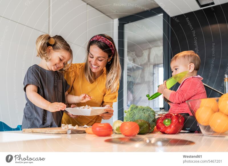 Lächelnde Mutter und Mädchen bereiten Essen zu, während die kleine Tochter auf der Kücheninsel sitzt Farbaufnahme Farbe Farbfoto Farbphoto Innenaufnahme