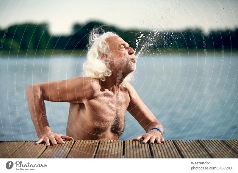 Älterer Mann mit weißen Haaren lehnt an Steg und spritzt mit Wasser Wasserspritzer Spritzer Wasser spritzen See Seen Senior ältere Männer älterer Mann Senioren