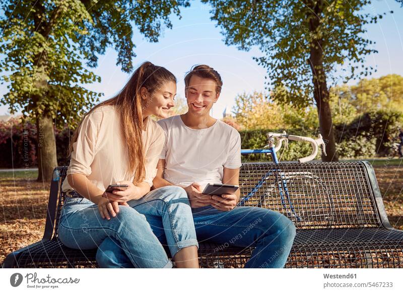 Junges Paar im Park sitzt auf einer Bank mit Handy und Tablet Sitzbänke Bänke Sitzbank Tablet Computer Tablet-PC Tablet PC iPad Tablet-Computer Pärchen Paare