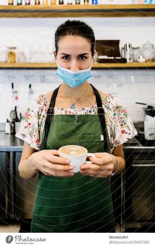 Junge Frau mit Gesichtsmaske und Schürze mit Kaffeetasse steht im Café Farbaufnahme Farbe Farbfoto Farbphoto Innenaufnahme Innenaufnahmen innen drinnen Tag