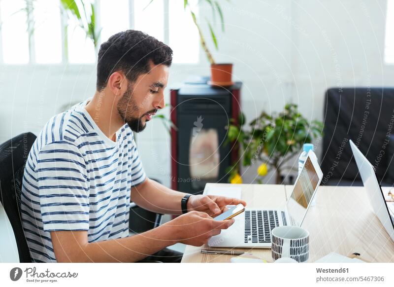 Junger Mann benutzt Laptop und Handy am Schreibtisch im Büro Notebook Laptops Notebooks Office Büros Arbeitstisch Schreibtische Mobiltelefon Handies Handys