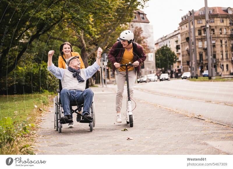Älterer Mann im Rollstuhl vergnügt sich mit seinen erwachsenen Enkelkindern im Freien Leute Menschen People Person Personen Europäisch Kaukasier kaukasisch