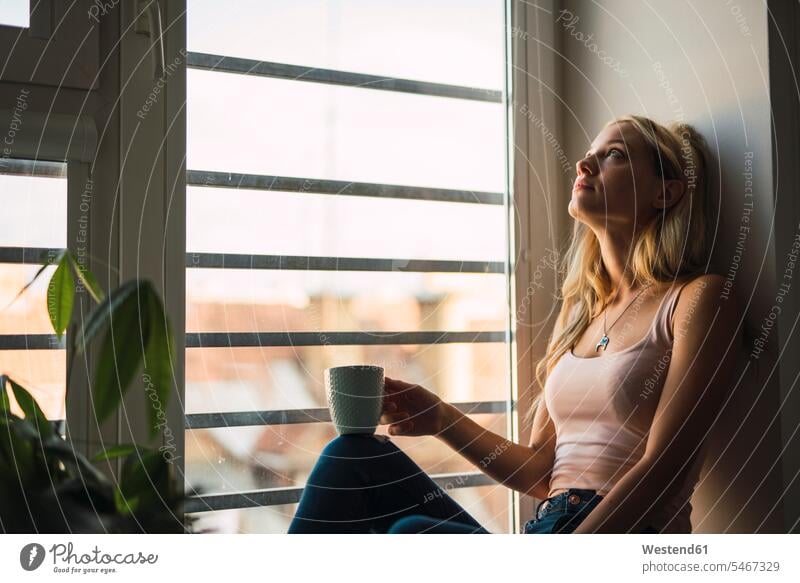 Blonde junge Frau hält Kaffeebecher sitzt in Fensterbank Becher blond blonde Haare blondes Haar sitzen sitzend schauen sehend Fensterbrett Fenstersims