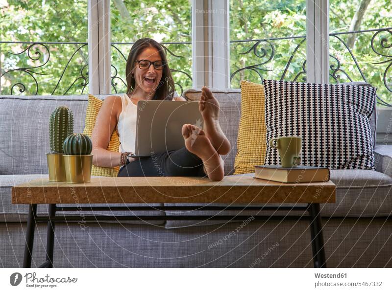 Fröhliche Frau benutzt Laptop, während sie im Wohnzimmer auf dem Sofa sitzt Farbaufnahme Farbe Farbfoto Farbphoto Innenaufnahme Innenaufnahmen innen drinnen Tag