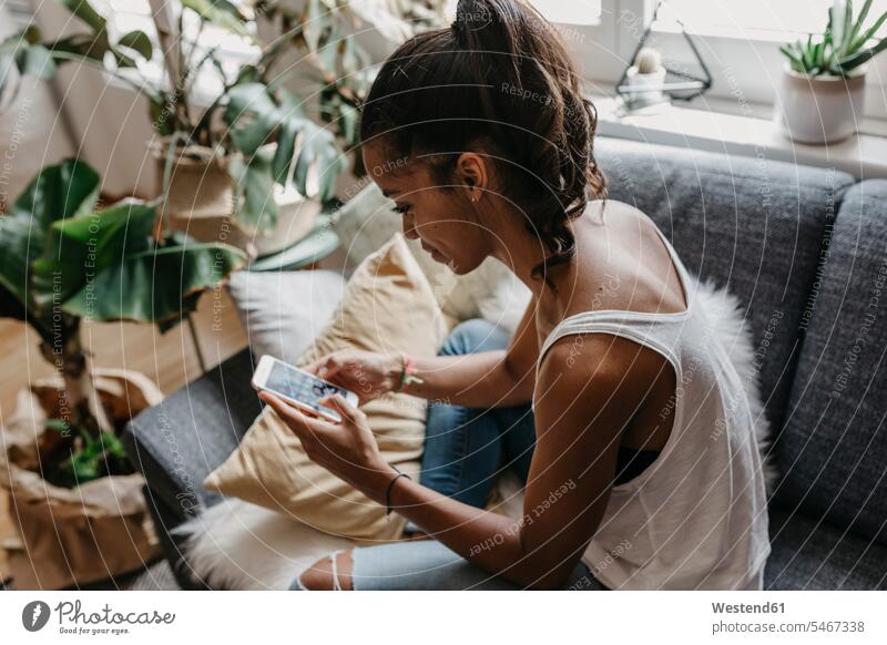 Junge Frau sitzt auf der Couch zu Hause mit Handy Zuhause daheim Sofa Couches Liege Sofas Smartphone iPhone Smartphones sitzen sitzend weiblich Frauen benutzen