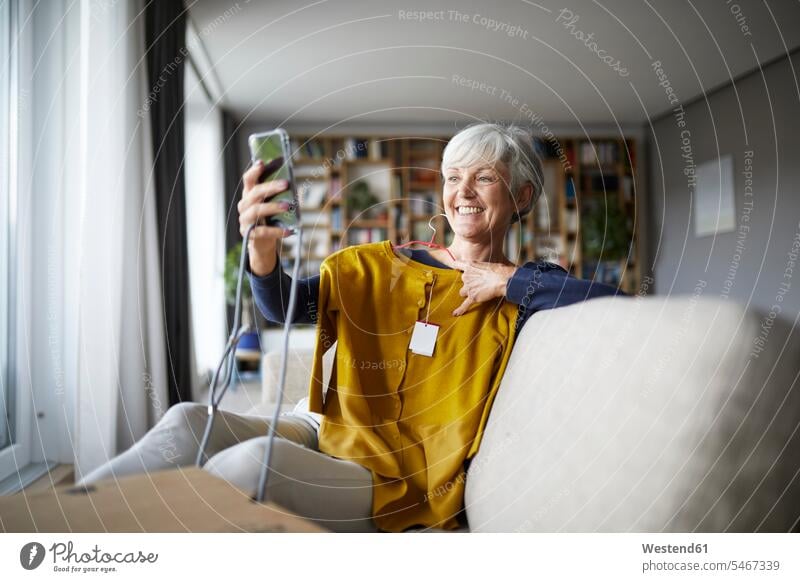 Lächelnde ältere Frau, die sich mit einer neuen Bluse auf einem Smartphone mit sich selbst beschäftigt, während sie zu Hause sitzt Farbaufnahme Farbe Farbfoto