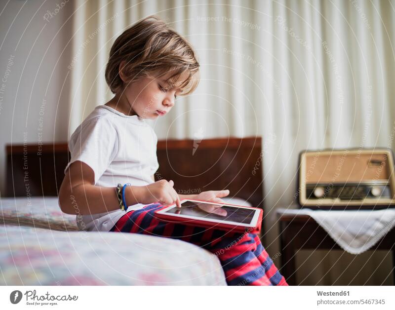 Junger Junge sitzt auf dem Bett und benutzt ein Tablet Buben Knabe Jungen Knaben männlich Tablet Computer Tablet-PC Tablet PC iPad Tablet-Computer Betten sitzen