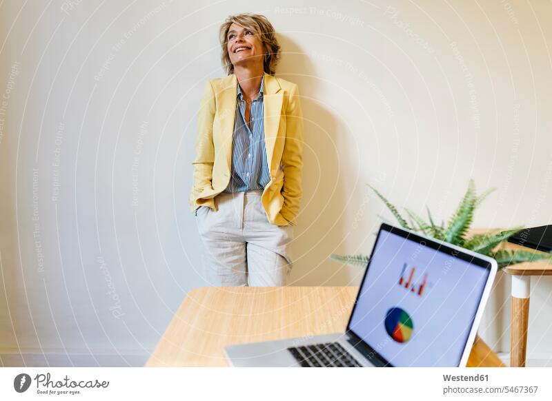 Lächelnde Geschäftsfrau mit Händen in den Taschen, die aufschaut, während sie im Büro an der Wand steht Farbaufnahme Farbe Farbfoto Farbphoto Innenaufnahme