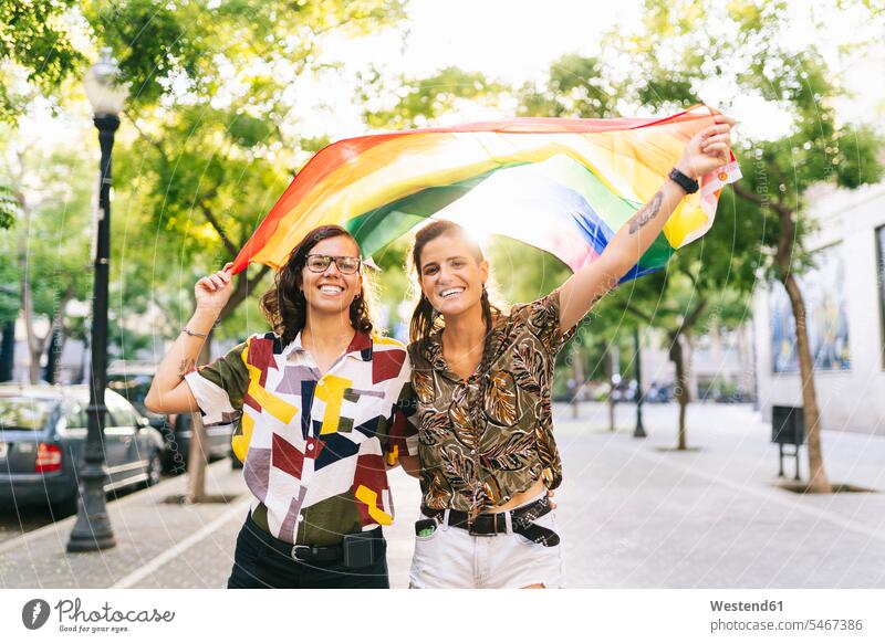 Glückliches lesbisches Paar schwenkt Regenbogenfahne, während es auf einem Fußweg in der Stadt steht Farbaufnahme Farbe Farbfoto Farbphoto Außenaufnahme außen