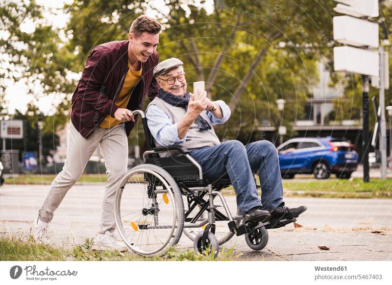 Lächelnder junger Mann schiebt glücklichen älteren Mann mit Smartphone im Rollstuhl Leute Menschen People Person Personen Europäisch Kaukasier kaukasisch 2