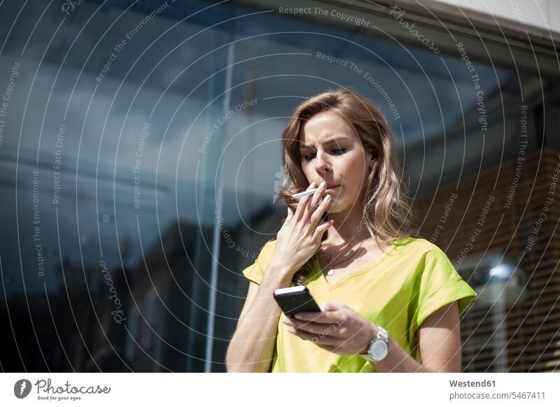 Junge Unternehmerin benutzt Smartphone, während sie an einem sonnigen Tag vor einem Bürogebäude raucht Farbaufnahme Farbe Farbfoto Farbphoto Außenaufnahme außen