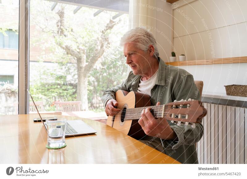 Pensionierter älterer Mann lernt Gitarre spielen durch Online-Tutorials am Laptop zu Hause Farbaufnahme Farbe Farbfoto Farbphoto Innenaufnahme Innenaufnahmen
