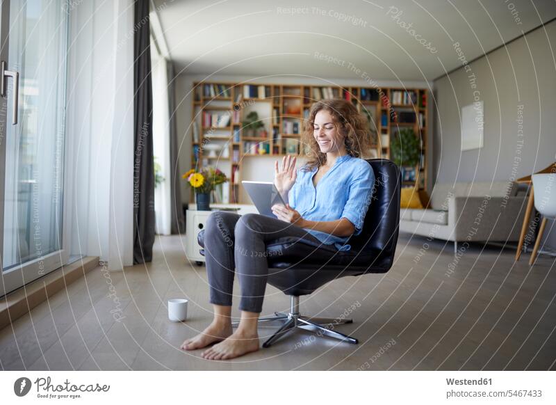 Lächelnde Frau winkt bei Videoanruf auf digitalem Tablett mit der Hand, während sie zu Hause auf einem Stuhl sitzt Farbaufnahme Farbe Farbfoto Farbphoto