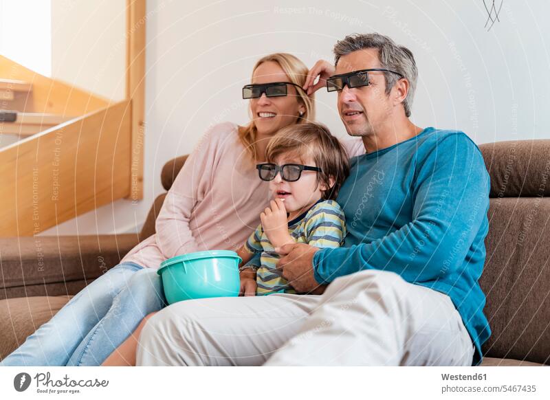 Vater, Mutter und Sohn tragen zu Hause auf der Couch eine 3D-Brille und sehen fern Schale Schalen Schälchen Schüsseln Couches Liege Sofas sehend sitzend sitzt