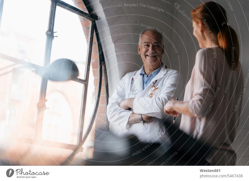 Lächelnder Oberarzt im Gespräch mit Frau am Fenster in einer Arztpraxis Arztpraxen Praxis Senior ältere Männer älterer Mann Senioren Doktoren Ärzte sprechen