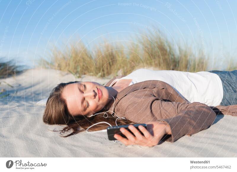 Porträt einer Frau, die auf einer Stranddüne liegt und mit Kopfhörern und Smartphone Musik hört, Sardinien, Italien telefonieren Handies Handys Mobiltelefon