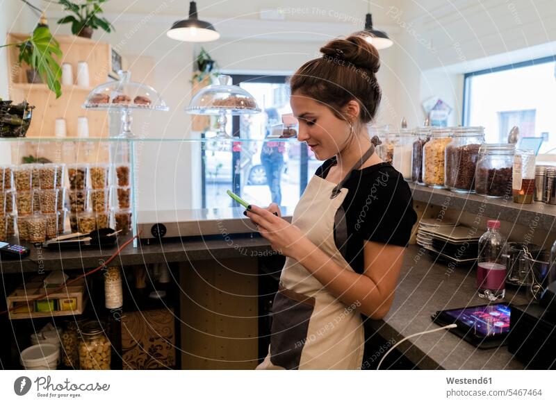 Junge Frau benutzt Handy hinter dem Tresen eines Cafés Job Berufe Berufstätigkeit Beschäftigung Jobs Beleuchtungen Lampen Leuchte Informationen