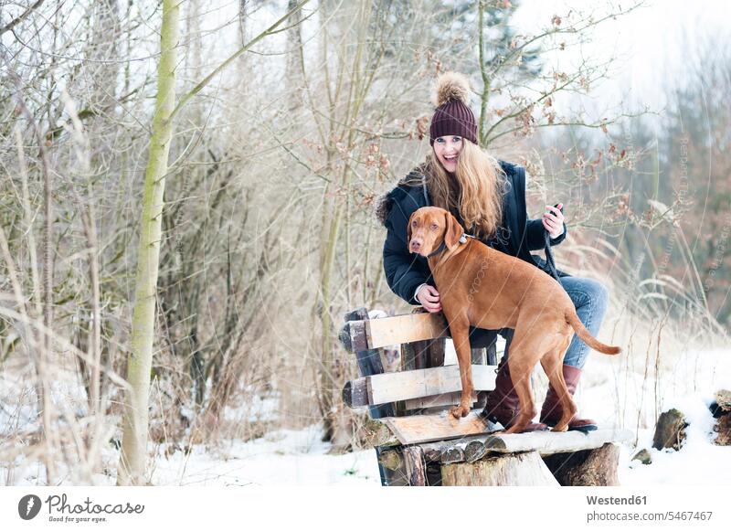 Fröhliche junge Frau sitzt im Winter auf einer Bank mit Hund vor kahlen Bäumen im Wald Freizeitbeschäftigung Muße Zeit Zeit haben ein Tier Einzelnes Tier 1