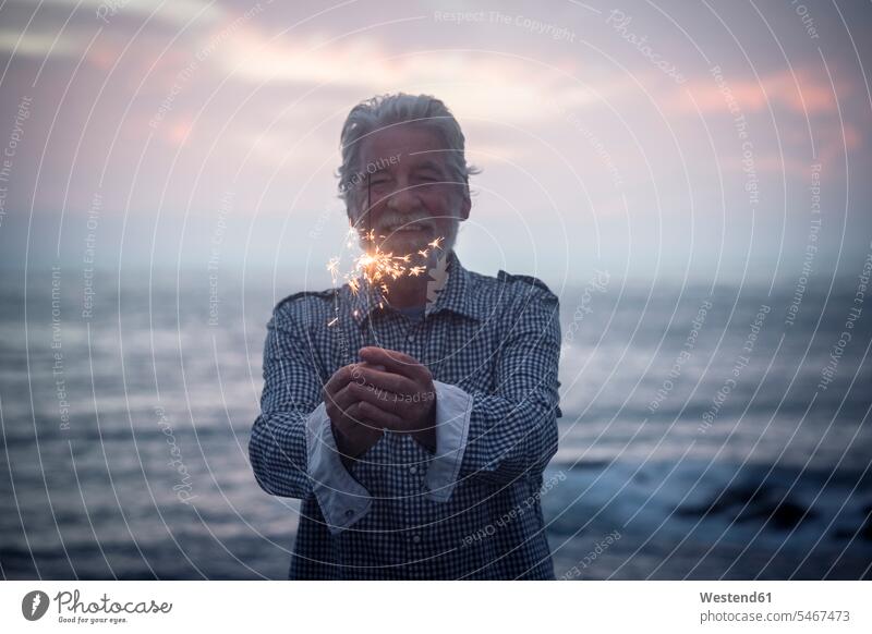 Glückliche ältere Mann steht vor dem Meer bei Sonnenuntergang hält Wunderkerze Spanien Vignettierung Abschattung Stimmungsvoller Himmel romantischer Himmel