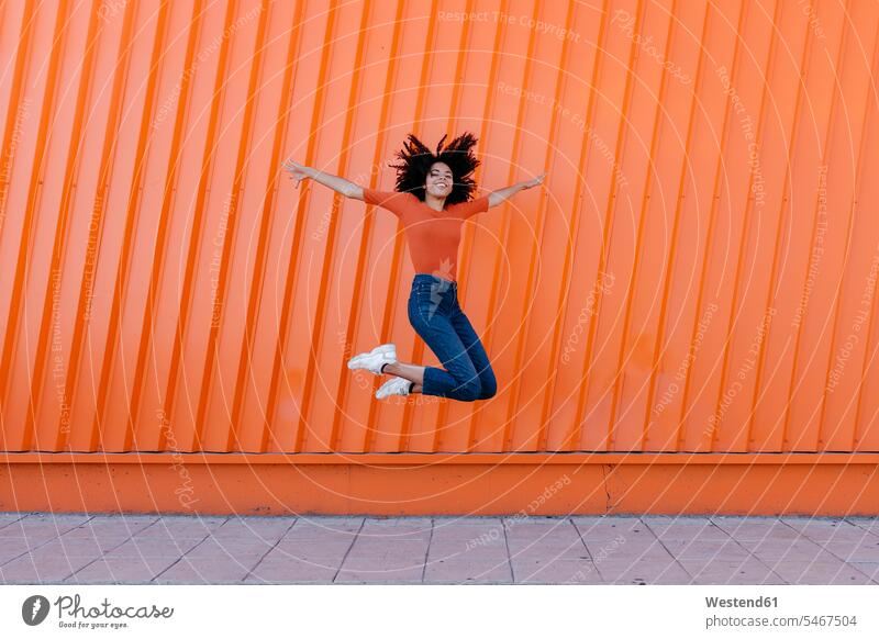 Glückliche junge Frau springt mit ausgestreckten Armen gegen orangefarbene Wand Farbaufnahme Farbe Farbfoto Farbphoto Außenaufnahme außen draußen im Freien Tag