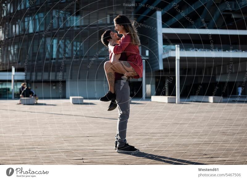 Junger Mann hebt glückliche Freundin auf Stadtplatz hoch hochheben Glück glücklich sein glücklichsein Platz Plätze Plaetze Paar Pärchen Paare Partnerschaft