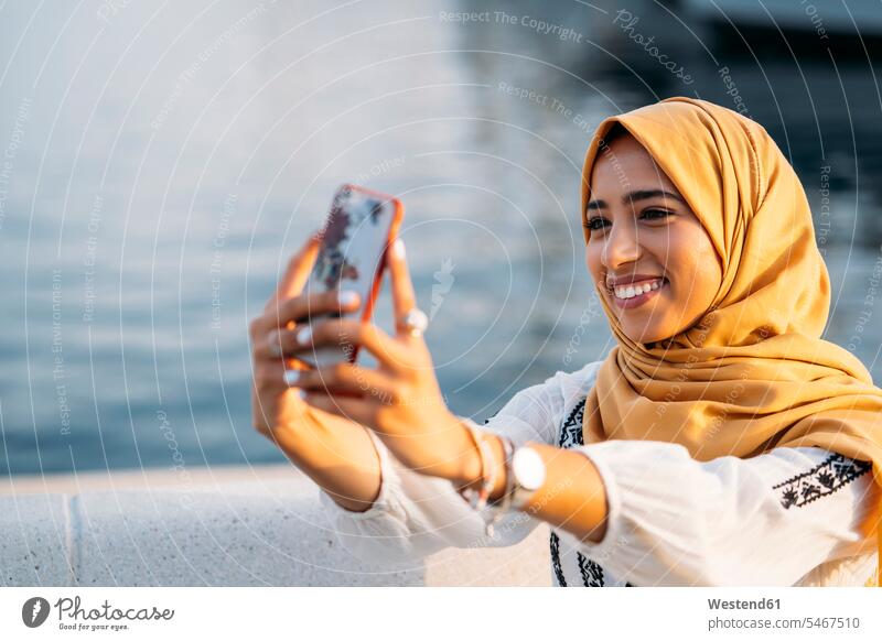 Junge muslimische Frau, die einen gelben Hidschab trägt, ein Smartphone benutzt und ein Selfie macht Leute Menschen People Person Personen Nordafrikanisch 1 Ein