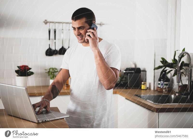 Lächelnder junger Mann mit Laptop und Handy in der Küche zu Hause Männer männlich telefonieren anrufen Anruf telephonieren lächeln Zuhause daheim Mobiltelefon