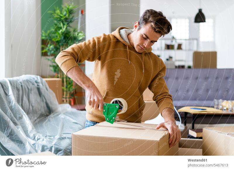 Junger Mann beim Anbringen von Klebeband auf Karton beim Umzug in ein neues Haus Farbaufnahme Farbe Farbfoto Farbphoto Innenaufnahme Innenaufnahmen innen