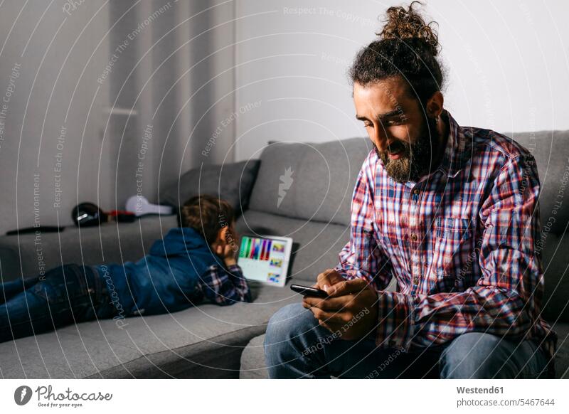 Vater sitzt auf der Couch mit Blick auf Smartphone, während sein Sohn mit digitalen Tablet im Hintergrund Couches Liege Sofas benützen nutzen sehen sehend