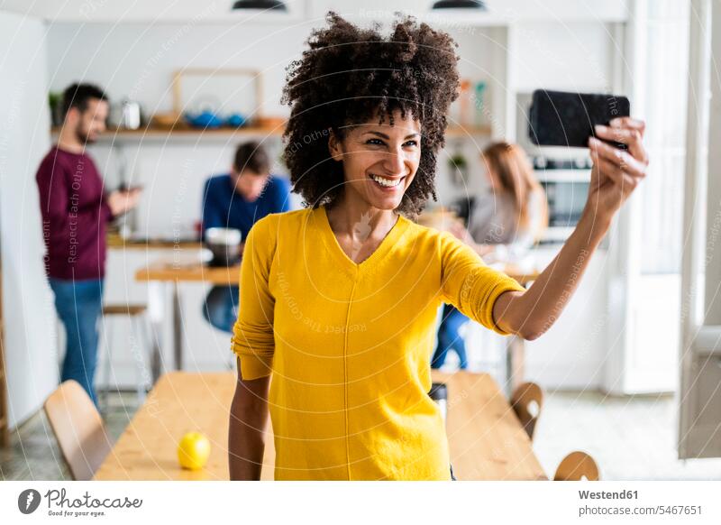 Glückliche Frau nimmt ein Selfie zu Hause mit Freunden im Hintergrund Leute Menschen People Person Personen Europäisch Kaukasier kaukasisch Afrikanisch
