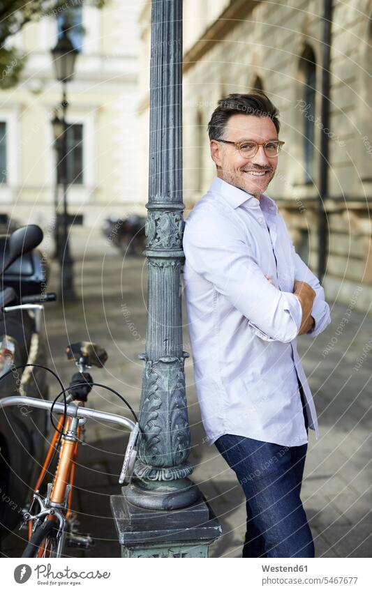 Porträt von lächelnden Geschäftsmann mit Fahrrad lehnt gegen Laternenpfahl in der Stadt Portrait Porträts Portraits staedtisch städtisch Businessmann