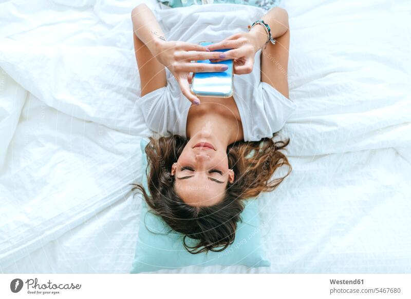 Draufsicht auf eine junge Frau, die im Bett liegt und ein Mobiltelefon benutzt Leute Menschen People Person Personen Europäisch Kaukasier kaukasisch 1 Ein