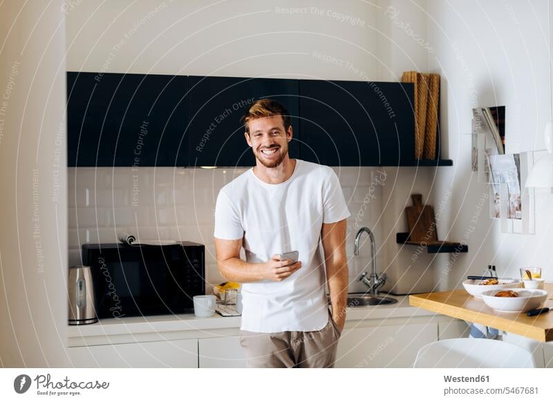 Porträt eines glücklichen jungen Mannes mit Handy in der Küche Leute Menschen People Person Personen Alleinstehende Alleinstehender Singles Unverheiratete