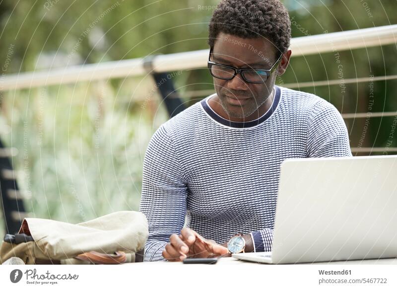 Männlicher Fachmann benutzt Mobiltelefon, während er mit Laptop am Tisch sitzt Farbaufnahme Farbe Farbfoto Farbphoto Außenaufnahme außen draußen im Freien Tag